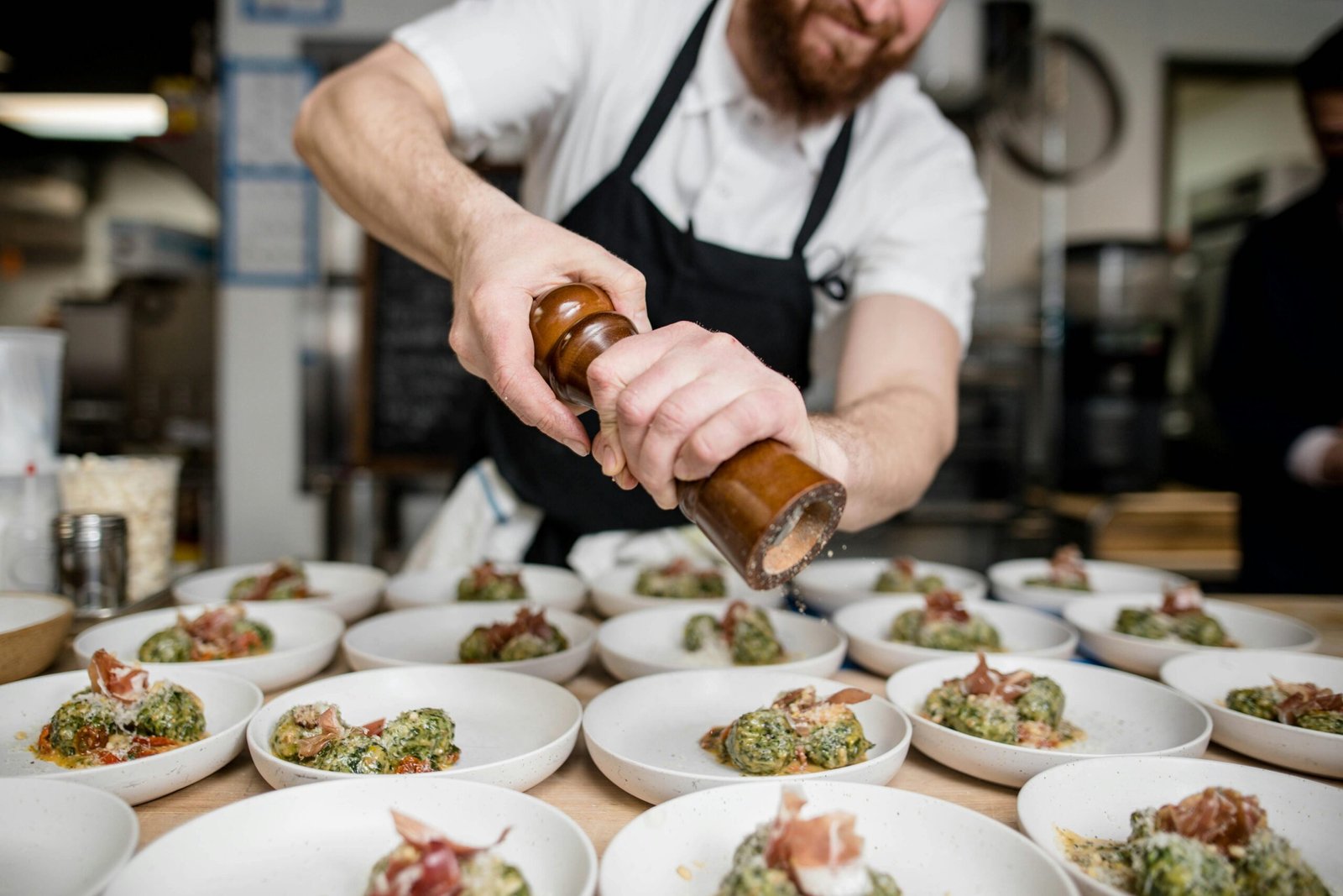 Bearded chef seasoning gourmet dishes with pepper in a busy restaurant kitchen.